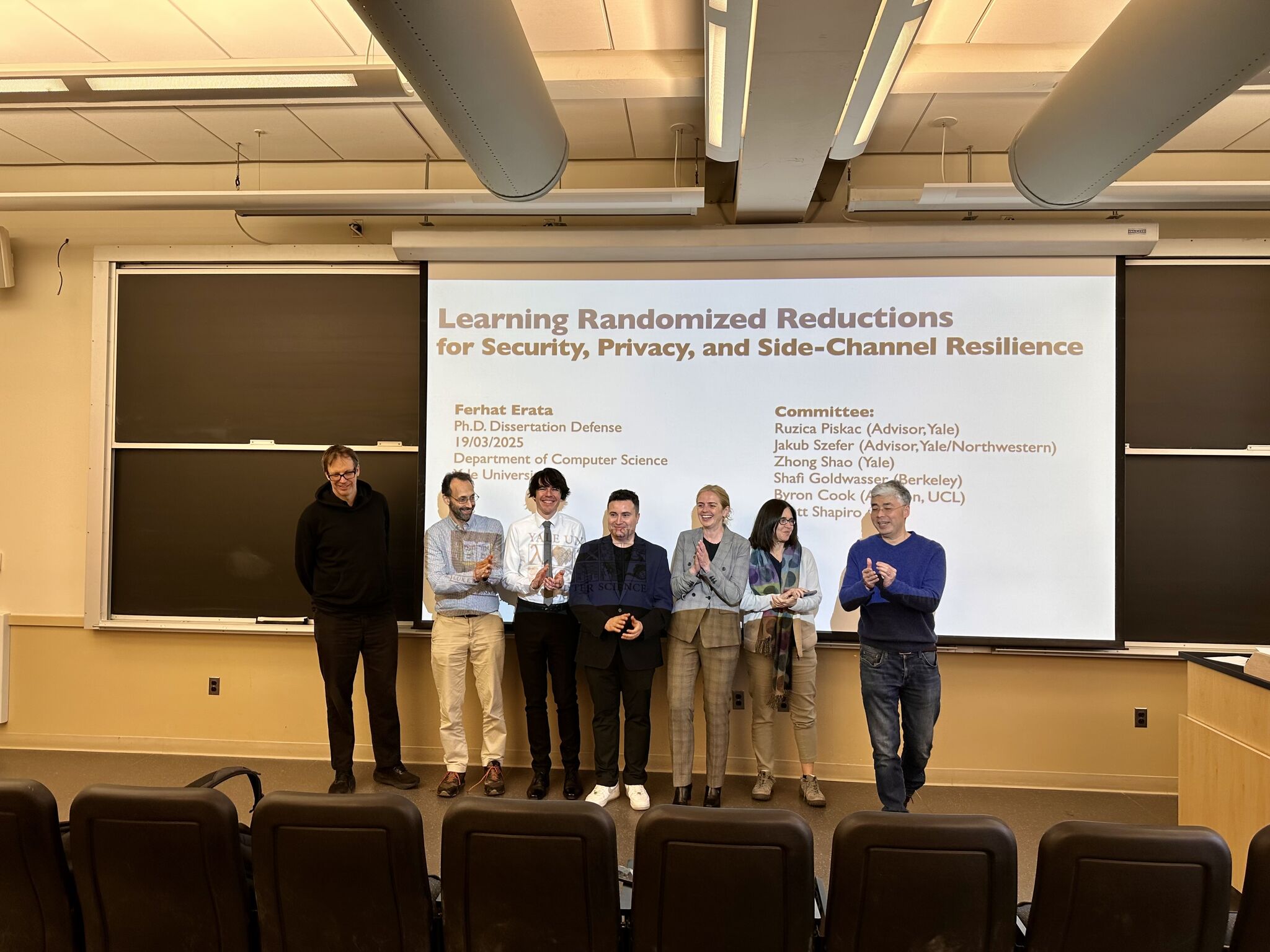 Ferhat Erata with his PhD thesis committee after defending at Yale, standing in front of a projected title slide